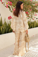 Woman in a beige outfit standing outdoors with flowers and greenery in the background