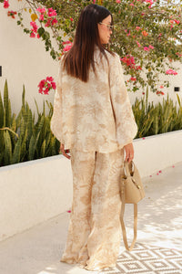 Woman in a beige outfit standing outdoors with flowers and greenery in the background