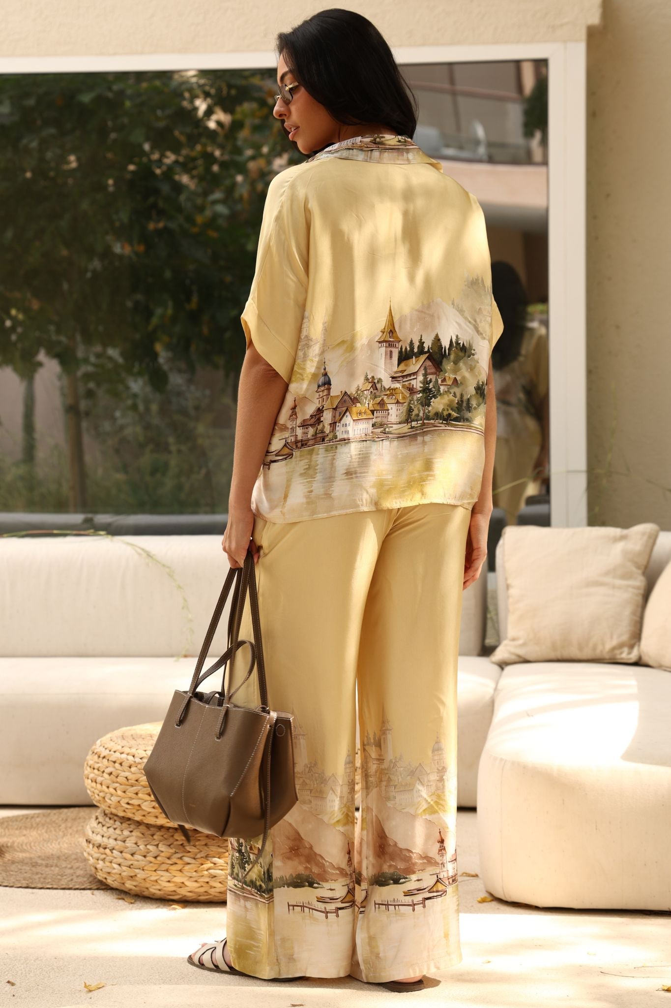 Woman in a yellow outfit with a scenic print, holding a brown handbag, standing in a living room.