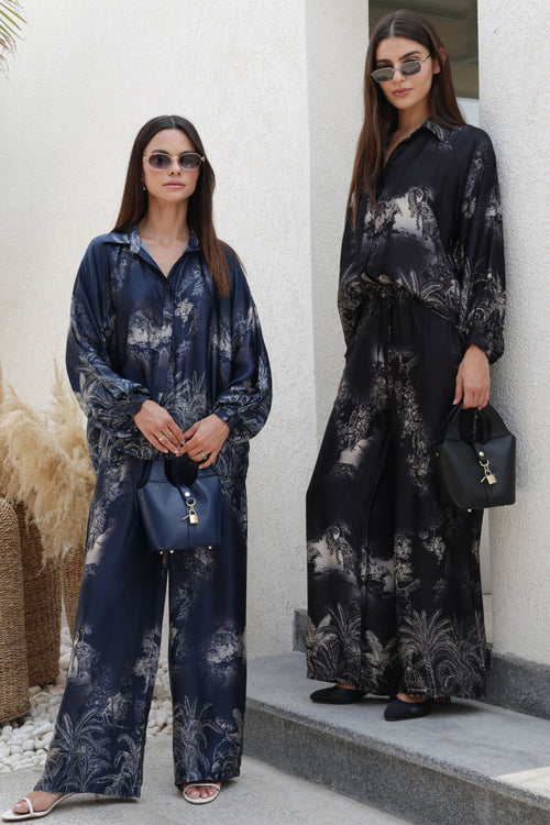 Two women wearing matching black and navy patterned outfits standing against a white wall.