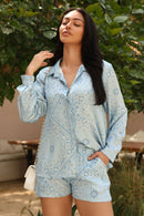 Woman wearing a light blue patterned outfit standing outdoors with greenery in the background