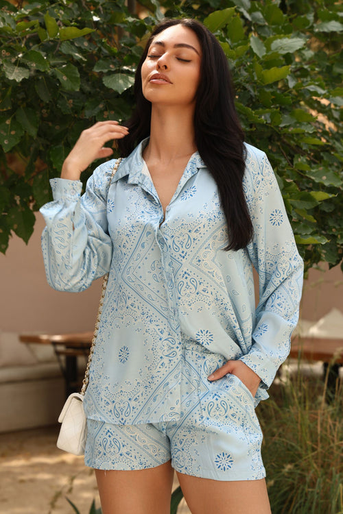 Woman wearing a light blue patterned outfit standing outdoors with greenery in the background