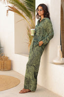 Woman in a green patterned outfit holding a cup, standing in a bright room with plants and decor.