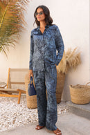 Woman in a blue patterned outfit standing in a sunlit outdoor setting with plants and a chair.