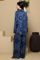 Woman wearing a blue patterned outfit standing in a room with beige walls and a dark vase.