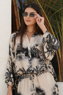 Woman wearing a black and white patterned outfit with sunglasses, standing against a white wall with plants.