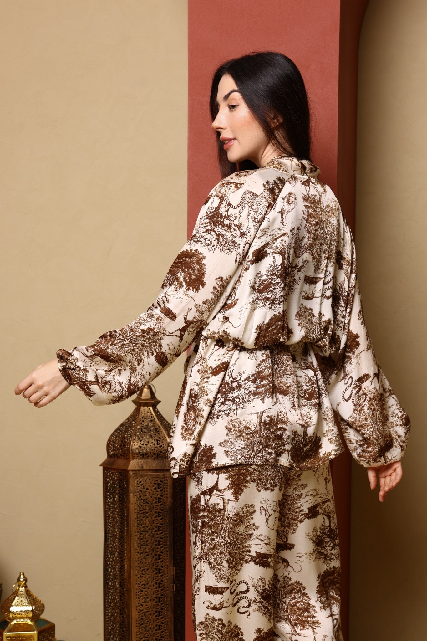 Woman wearing a patterned outfit standing next to a decorative lantern against a beige wall.