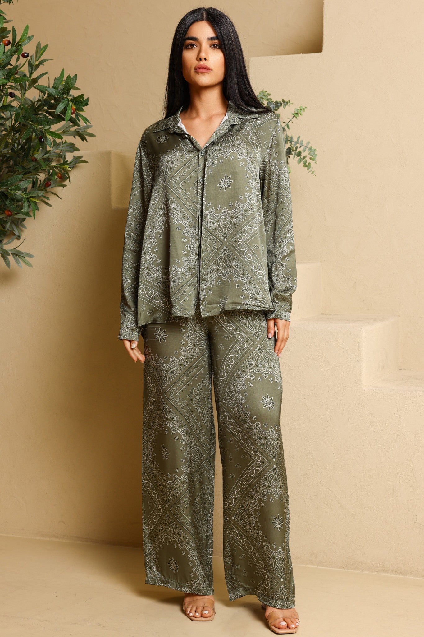 Woman wearing a green patterned outfit standing against a beige wall with decorative plants.