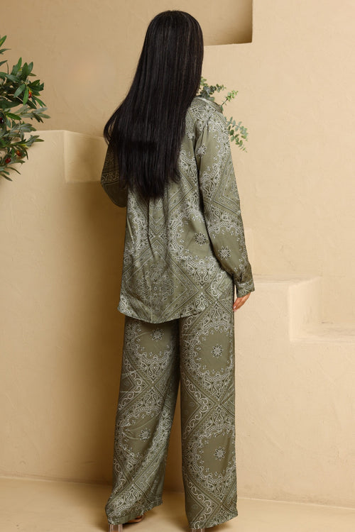 Person wearing a green patterned outfit standing against a beige wall with decorative plants.