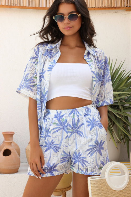Woman wearing a blue and white floral outfit with sunglasses, standing against a white wall with plants.