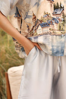 Person wearing a shirt with a scenic print of a village by a lake.
