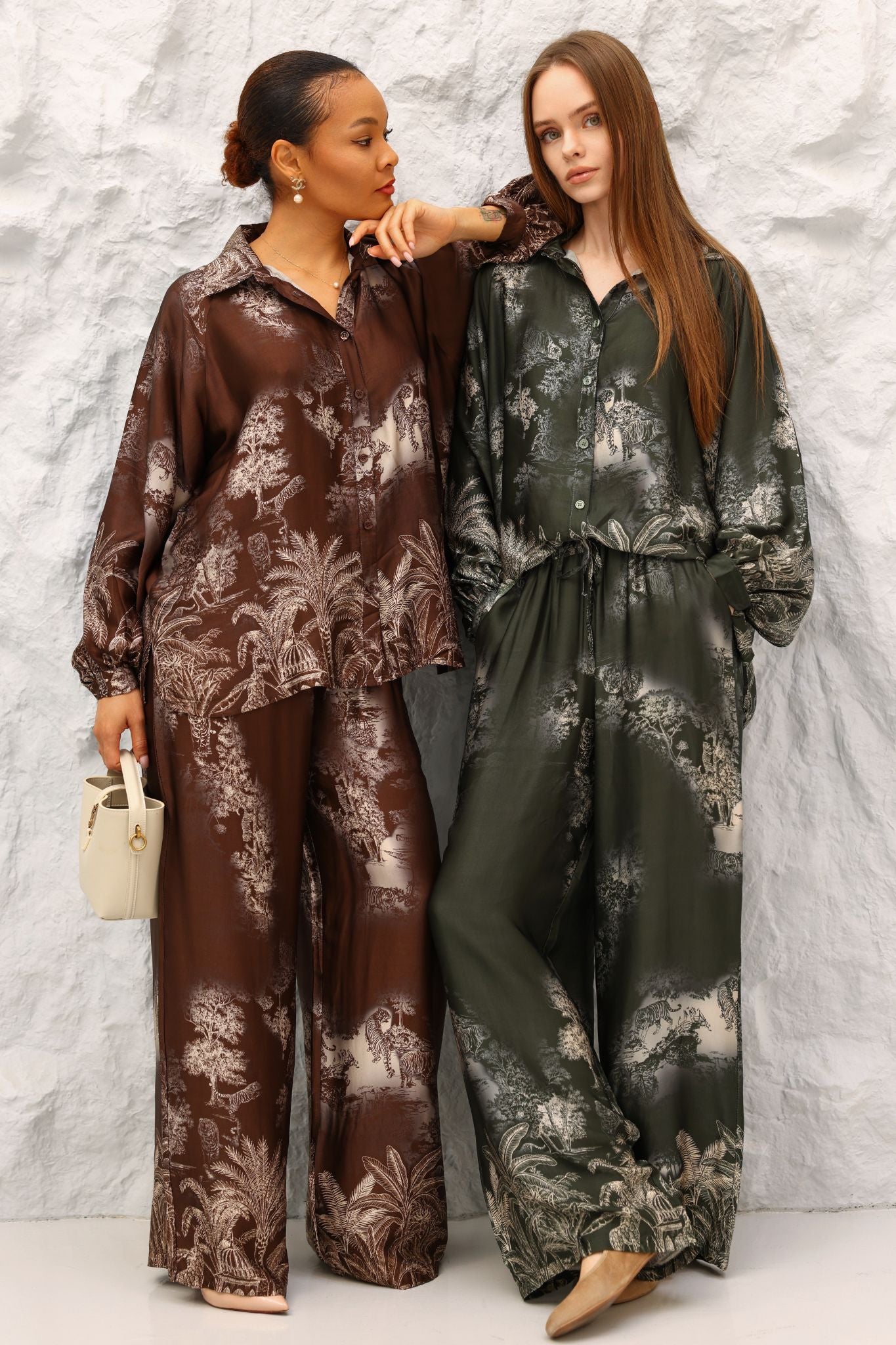 Two women wearing floral-patterned outfits standing against a textured white wall.