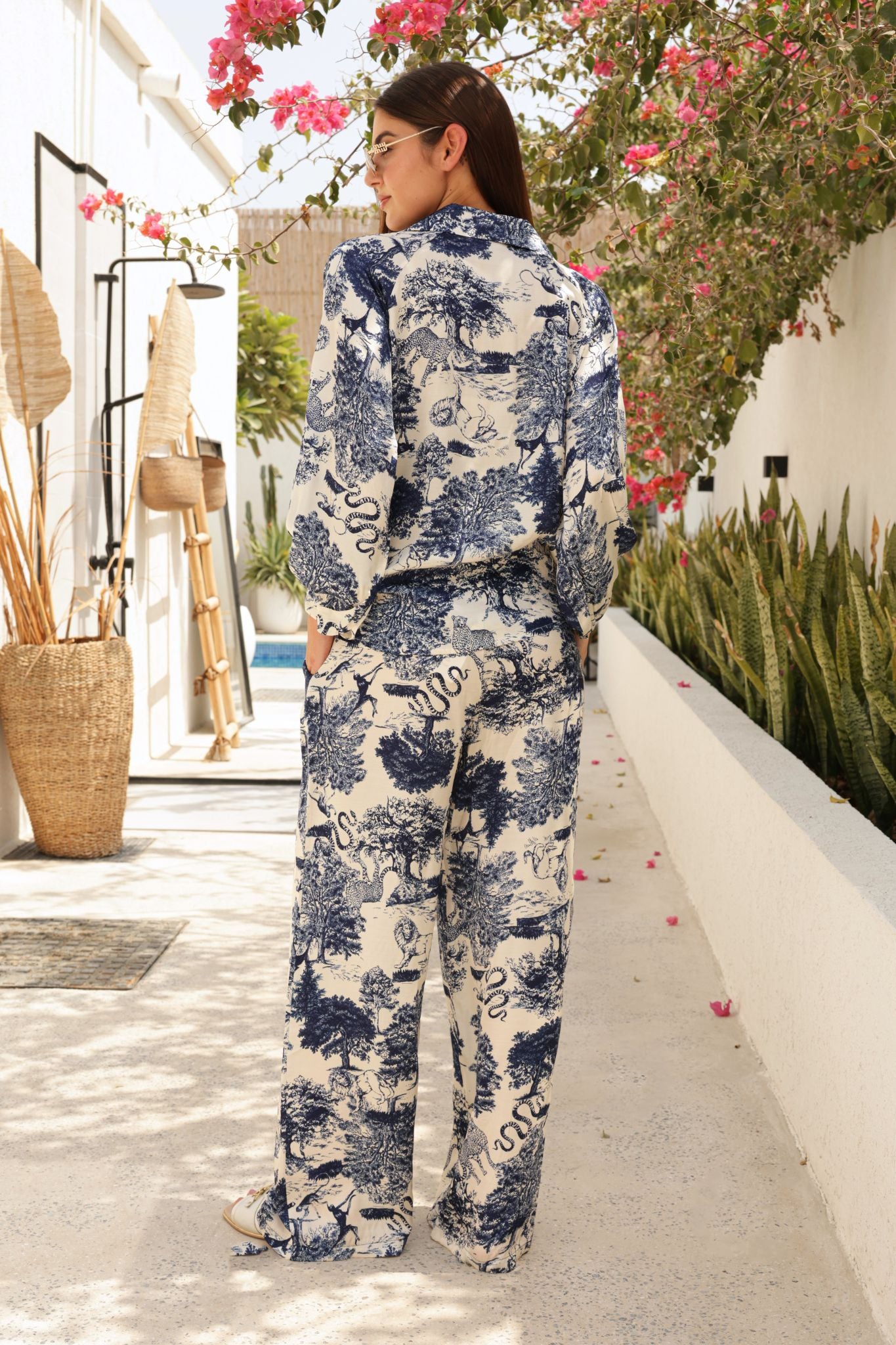 Woman wearing a blue and white floral outfit standing in a sunlit outdoor area with plants and flowers.