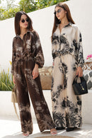 Two women in stylish outfits standing outdoors with a white wall and greenery in the background.