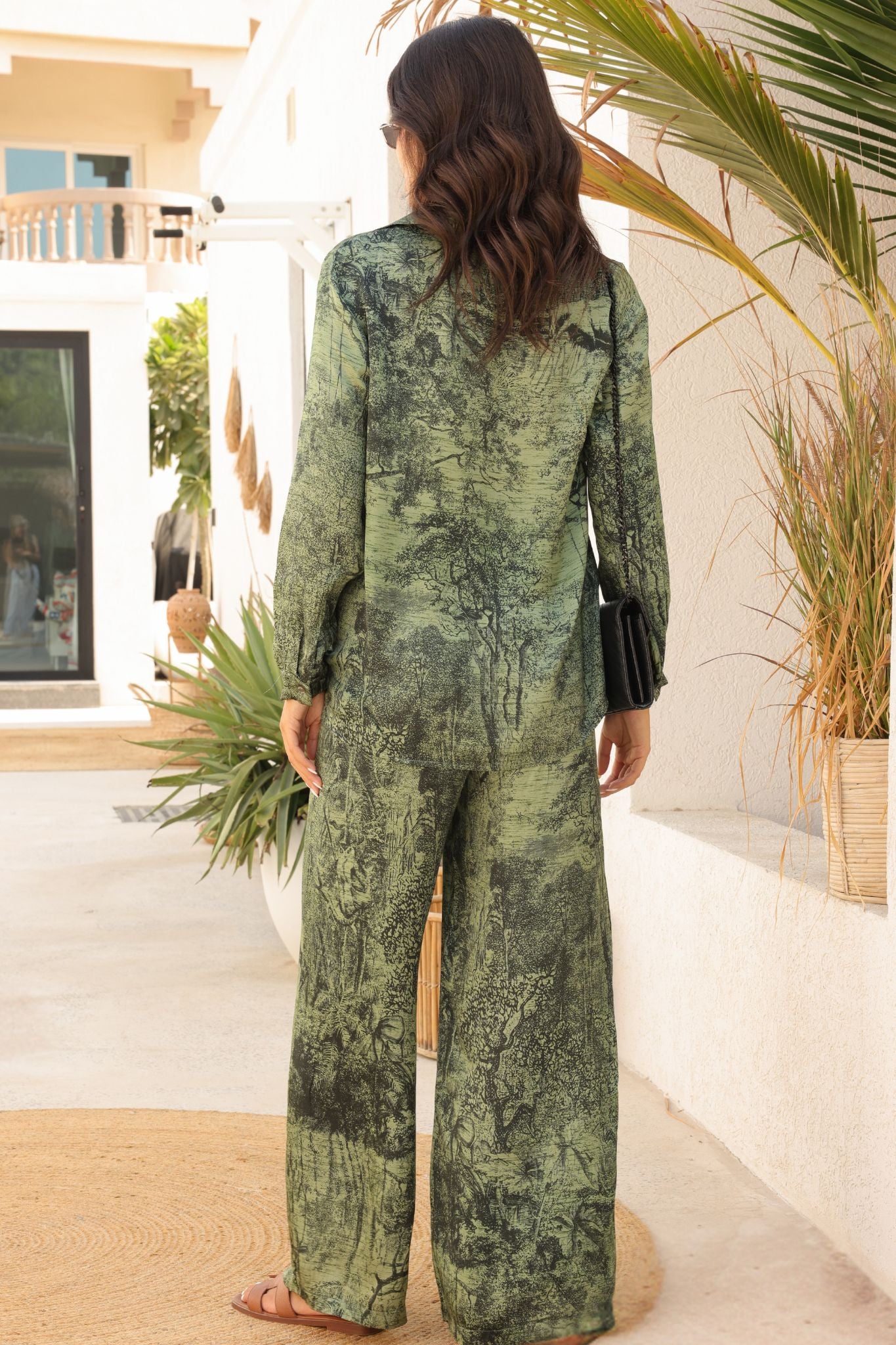 Woman in a green patterned outfit standing outdoors with plants and a building in the background