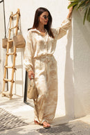 Woman in a beige outfit standing against a white wall with a plant.