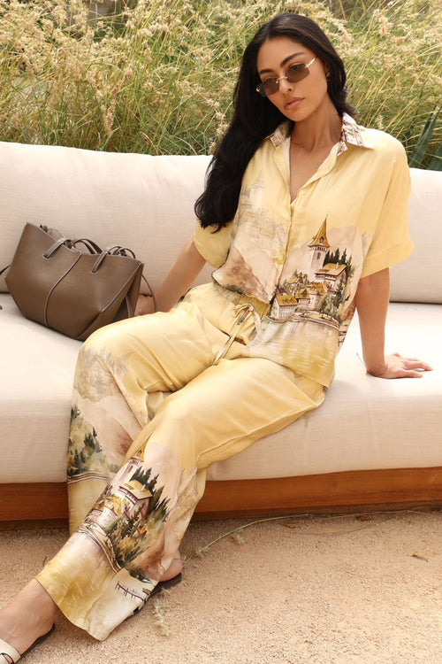 Woman sitting on a couch wearing a yellow outfit with floral patterns, holding a brown handbag.