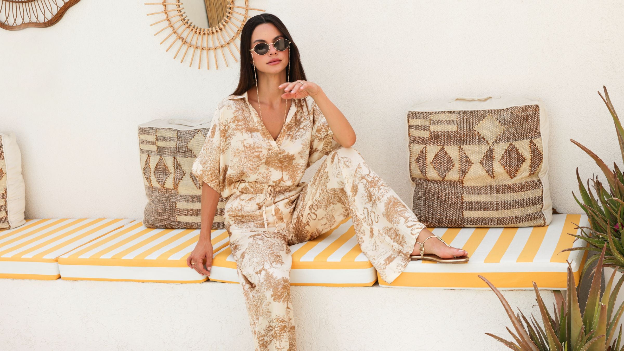 Woman in a gold outfit sitting on a bench with decorative pillows and plants.