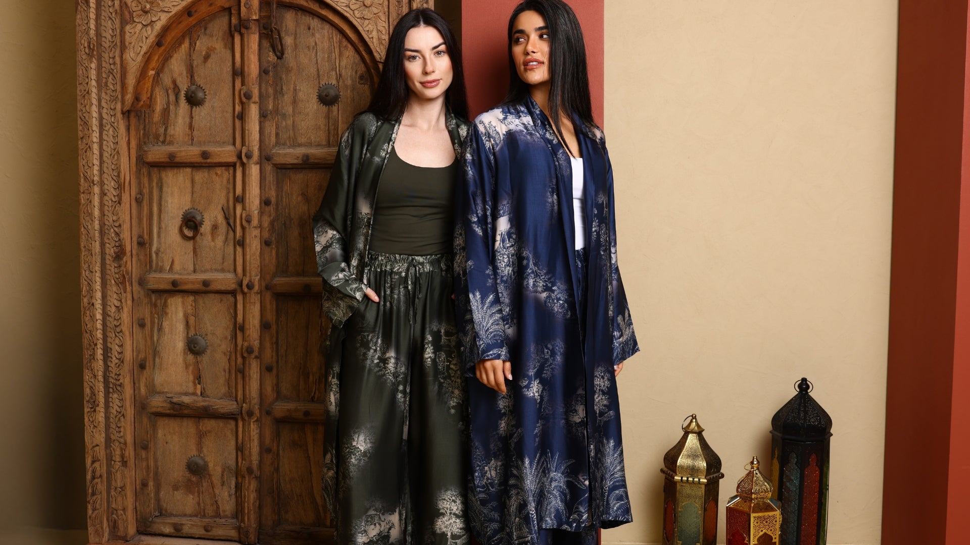 Two women wearing long, patterned robes standing in front of a wooden door.